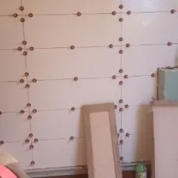 Białe płytki ścienne z drobnym wzorem, ułożone w regularną siatkę z użyciem krzyżyków dystansowych, widoczne narzędzia i materiały budowlane na podłodze, żółty sufit w tle.