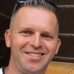 Portret uśmiechniętego mężczyzny z krótkimi włosami i okularami przeciwsłonecznymi na głowie, na tle brązowej ściany z drewnianą ramą.