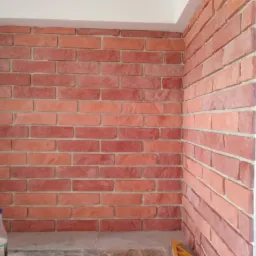 Ściana wyłożona cegiełkami klinkierowymi w odcieniach czerwieni i pomarańczy, widoczna butelka z płynem i fragment poziomicy.