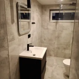 Nowoczesna łazienka z umywalką nablatową na czarnej szafce, lustrem z podświetleniem LED i szarą płytką imitującą beton na ścianach i podłodze, widoczny fragment kabiny prysznicowej.