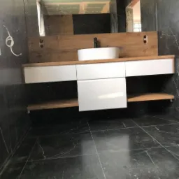 Łazienka w trakcie remontu: czarne marmurowe płytki, biała umywalka nablatowa na drewnianym blacie, widoczne kable i surowy sufit w odbiciu lustra.