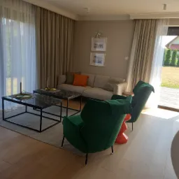 Jasne wnętrze salonu z parkietem, dwoma zielonymi fotelami na pomarańczowych nogach, szarą sofą z poduszkami i stolikami kawowymi z czarnymi blatami imitującymi marmur.