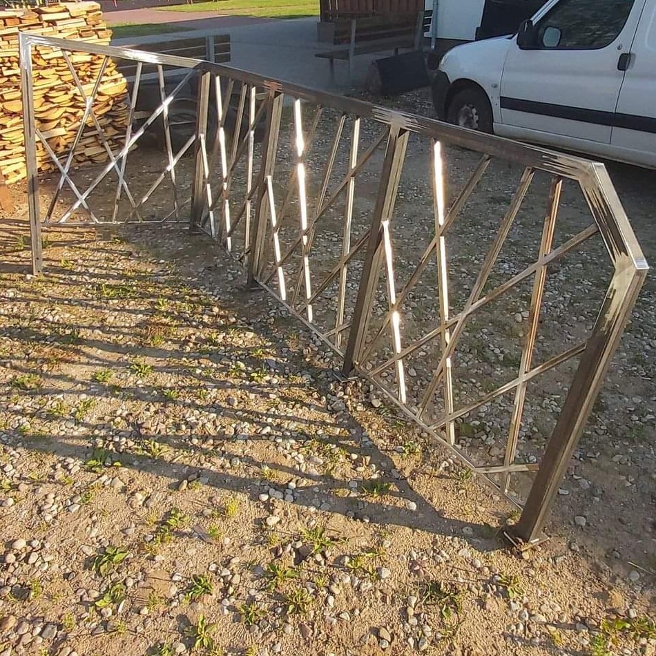 Surowa, metalowa konstrukcja ogrodzenia z ukośnymi prętami, leżąca na żwirowym podłożu w świetle słonecznym, z stosem drewna i białym samochodem w tle.