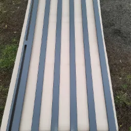 Element ogrodzenia metalowego w kolorze grafitowym, z pionowymi profilami i otworami montażowymi, leżący na kartonie na tle ziemi i trawy.