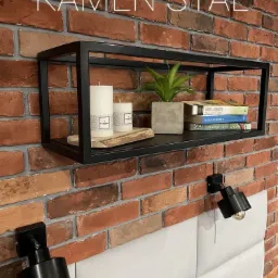 Czarna, prostokątna półka metalowa zamontowana na ceglanej ścianie, z dekoracjami w postaci świec, rośliny w betonowej doniczce i książek, oświetlona dwoma czarnymi lampkami ściennymi.