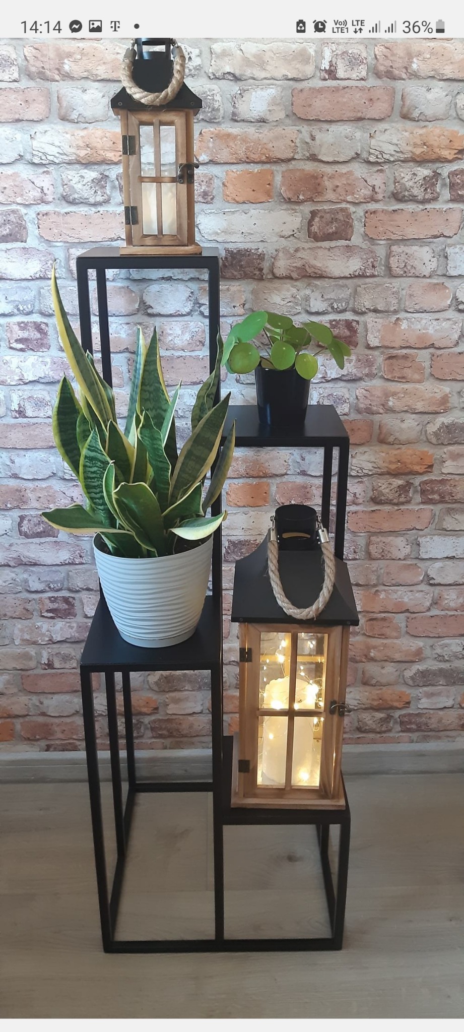 Metalowy regał schodkowy z trzema półkami, na których ustawiono rośliny doniczkowe i drewniane lampiony ze sznurkowymi uchwytami, na tle ceglanej ściany.