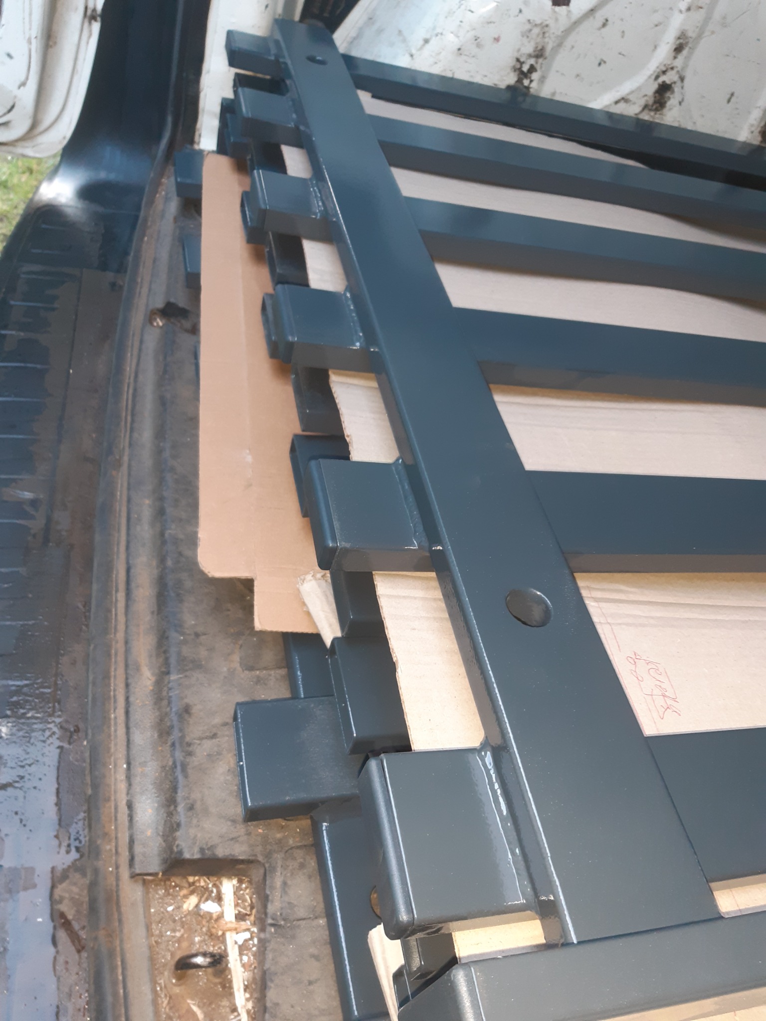 Elementy konstrukcyjne metalowe, prawdopodobnie do schodów, w trakcie transportu, zabezpieczone kartonem, widoczne spawy i otwory montażowe, szara farba.