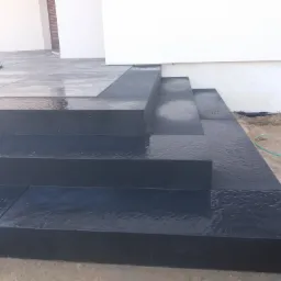 Nowoczesne, ciemne schody zewnętrzne z kamienia prowadzące do budynku z jasną elewacją, widoczne ślady wilgoci na powierzchni.