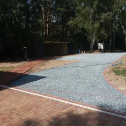 Utwardzony żwirem podjazd w trakcie budowy, obrzeża wykończone kostką, w tle zieleń i budynki gospodarcze.