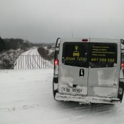 Tył białego busa A&W Taxi zaparkowanego na ośnieżonej drodze, z widocznymi numerem telefonu i ofertą usług: transport na lotniska, zakupy na telefon, wydarzenia okolicznościowe, pomoc na drodze.