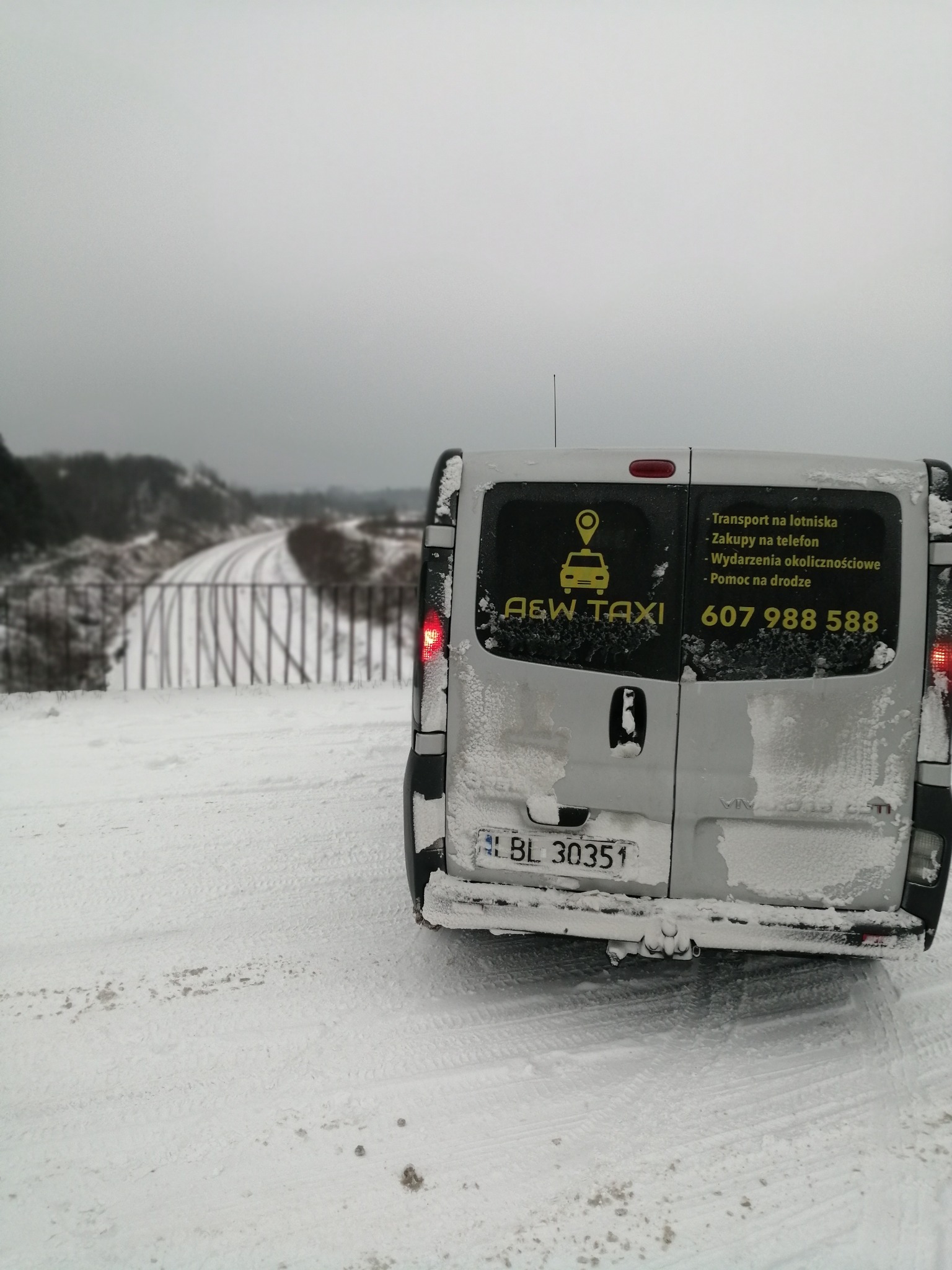 Tył białego busa A&W Taxi zaparkowanego na ośnieżonej drodze, z widocznymi numerem telefonu i ofertą usług: transport na lotniska, zakupy na telefon, wydarzenia okolicznościowe, pomoc na drodze.