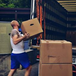 Home Warriors - Mężczyzna z tatuażem na ramieniu w białym podkoszulku i niebieskich spodenkach przenosi kartonowe pudło do wnętrza ciężarówki, widoczne pasy transportowe i inne kartony.