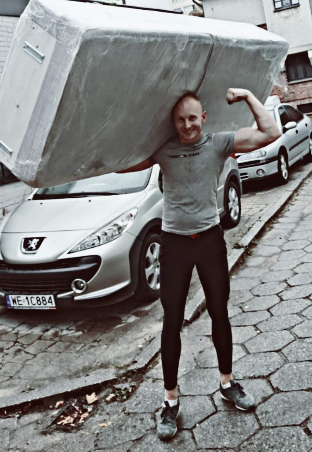 Uśmiechnięty mężczyzna z napiętym bicepsem, niosący na barkach duży, owinięty folią pakunek na tle dwóch samochodów osobowych na brukowanej ulicy.