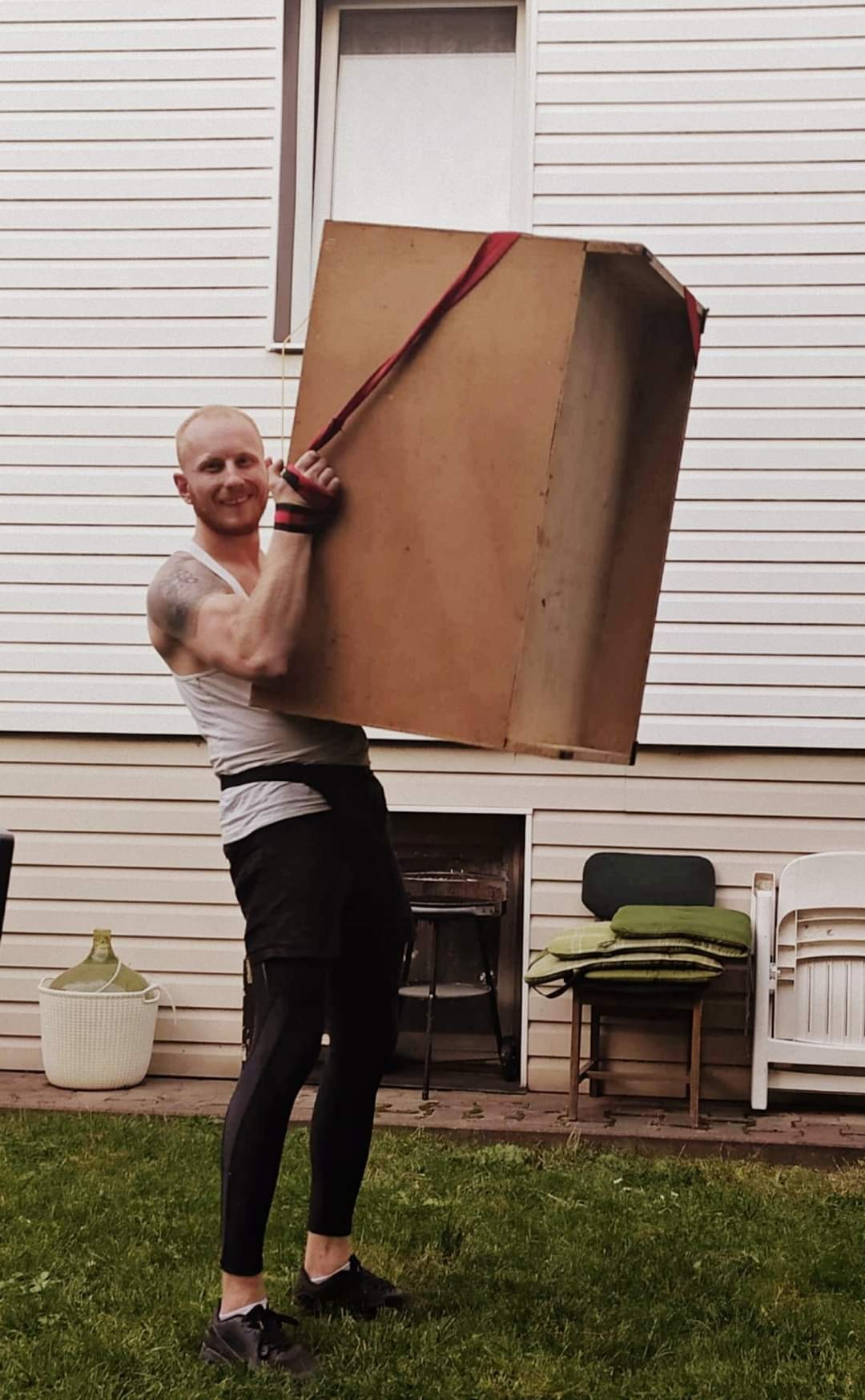 Uśmiechnięty mężczyzna z tatuażem na ramieniu, w sportowym stroju, przenosi dużą, kartonową skrzynię za pomocą czerwonych pasów transportowych na tle białego domu z oknem i zielonego trawnika.