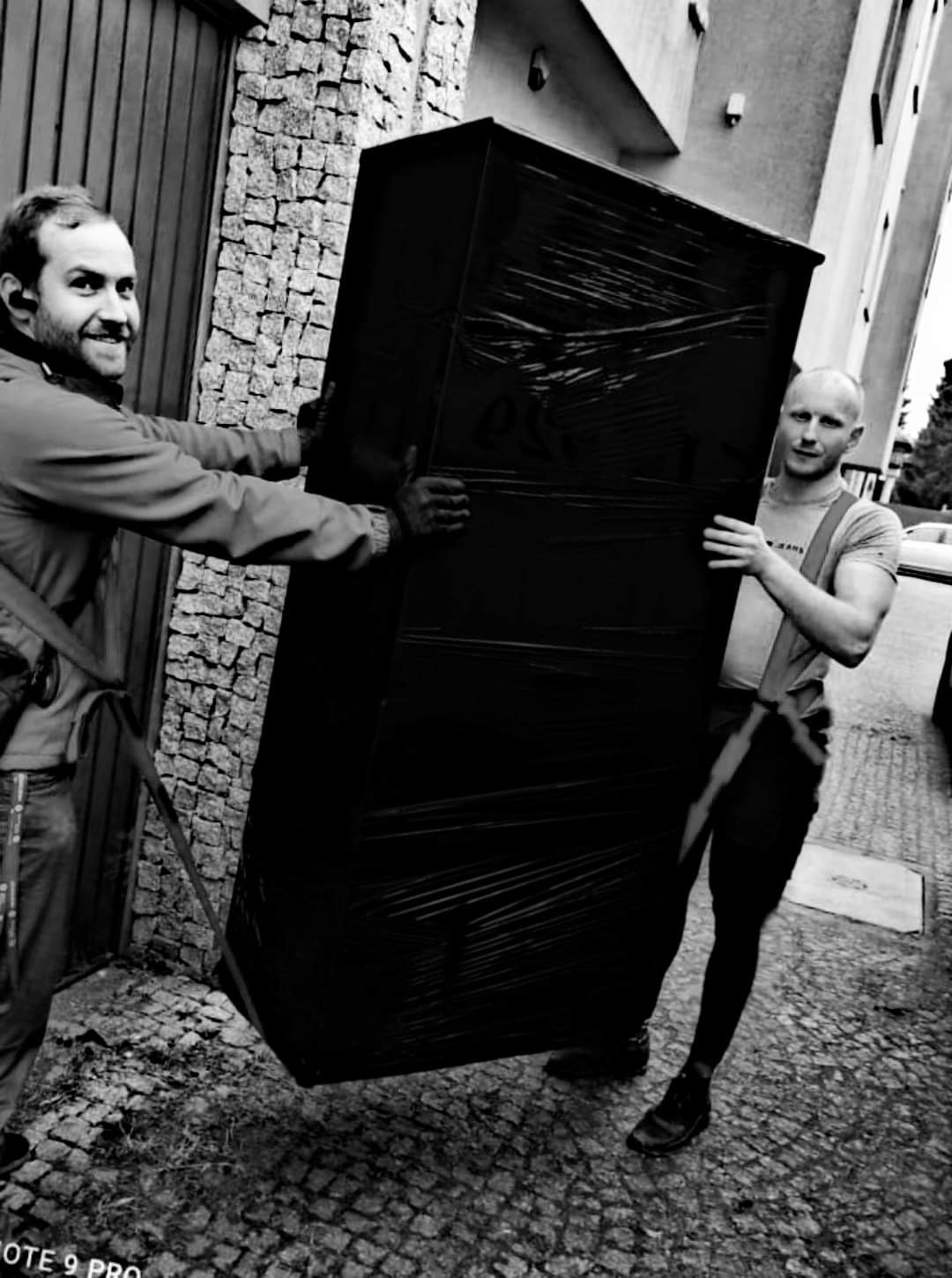 Dwóch mężczyzn w czarno-białym ujęciu przenosi dużą, owiniętą folią szafę na zewnątrz budynku z kamienną elewacją.