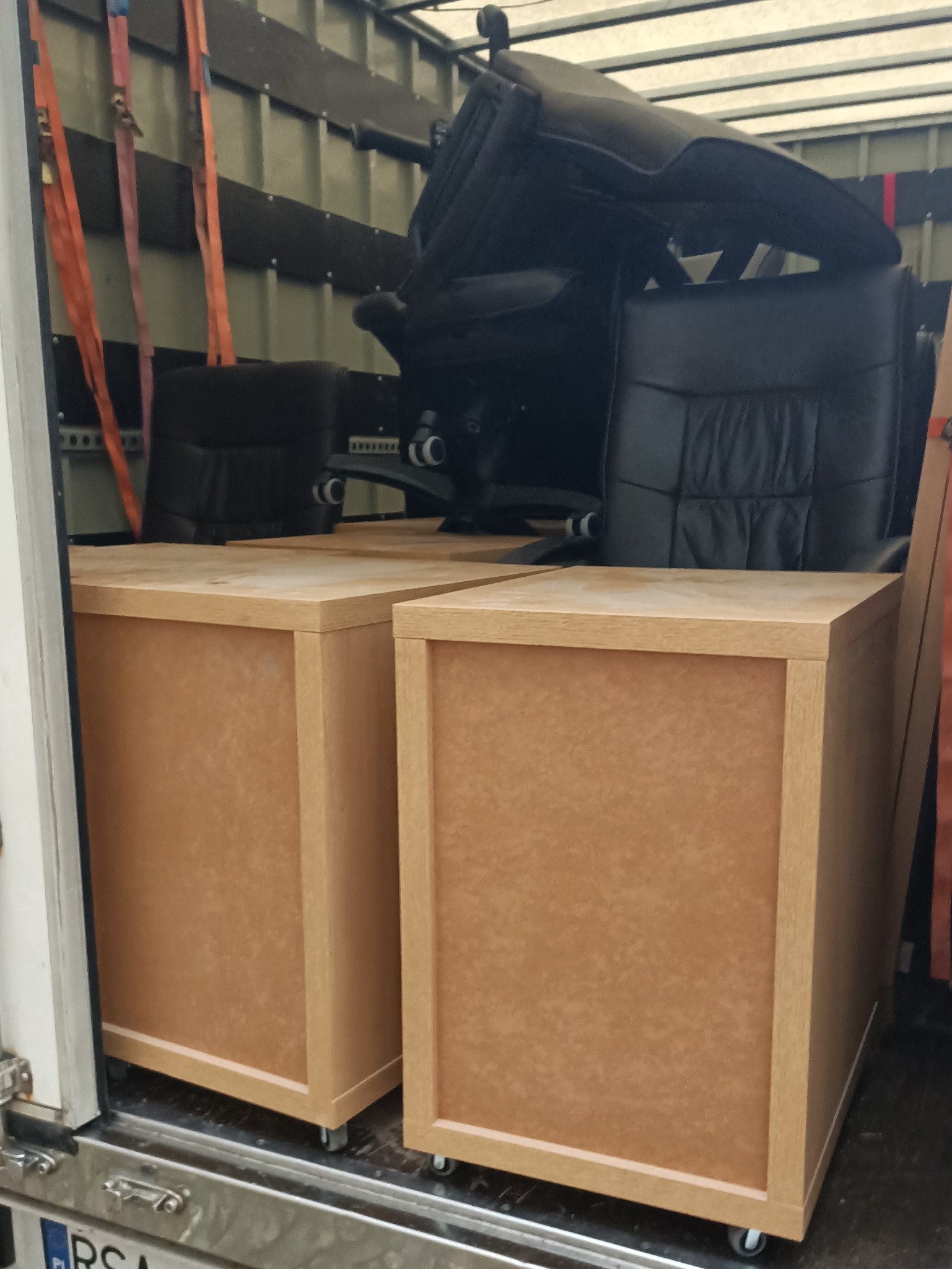 Dwa biurka z szufladami na kółkach i czarne skórzane krzesło biurowe zabezpieczone pasami wewnątrz furgonetki podczas transportu.