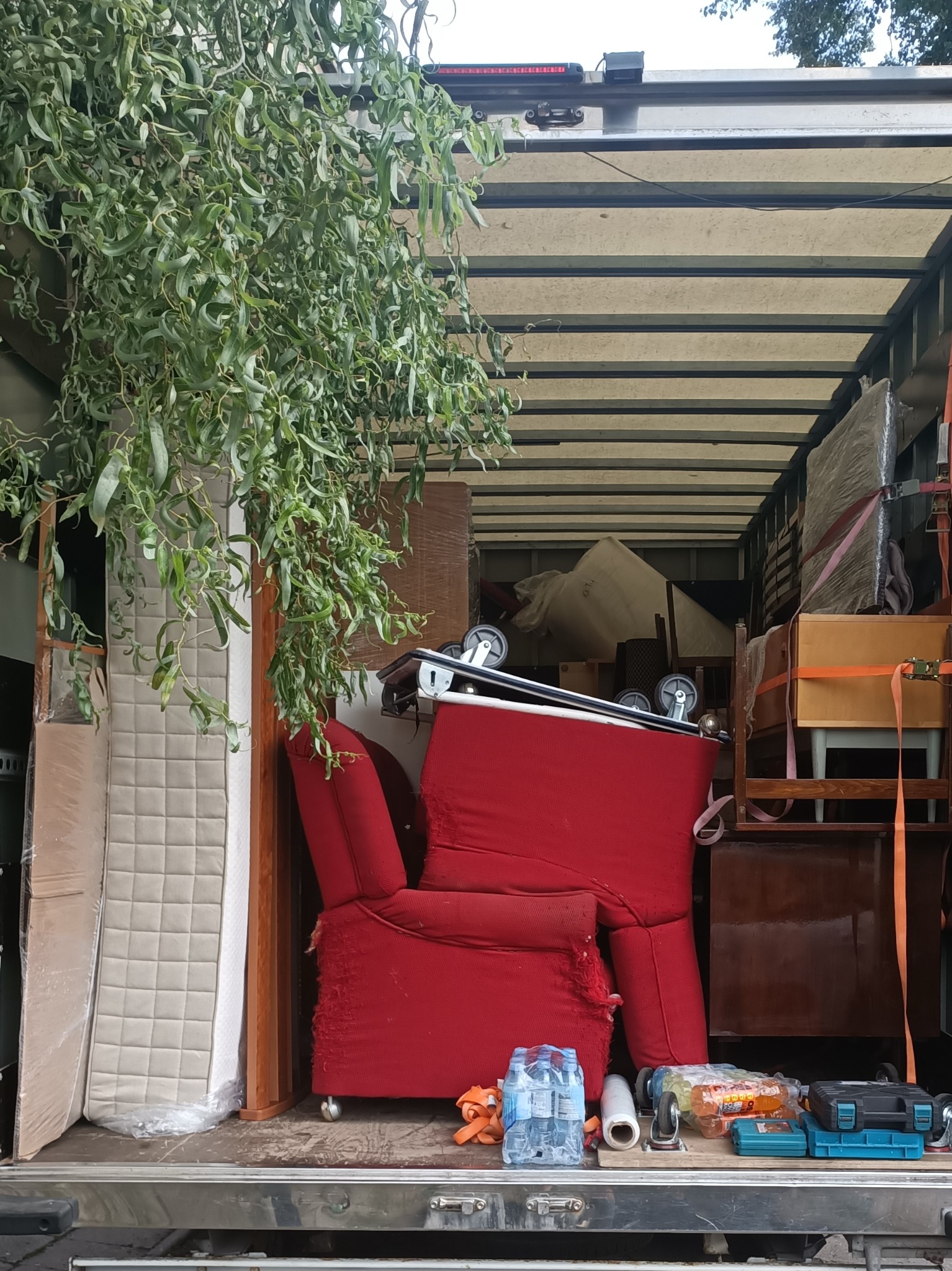 Wnętrze busa załadowane meblami i sprzętami podczas przeprowadzki, widoczny czerwony fotel z kółkami, materac, szafka i skrzynki z narzędziami, całość zabezpieczona pasami transportowymi.