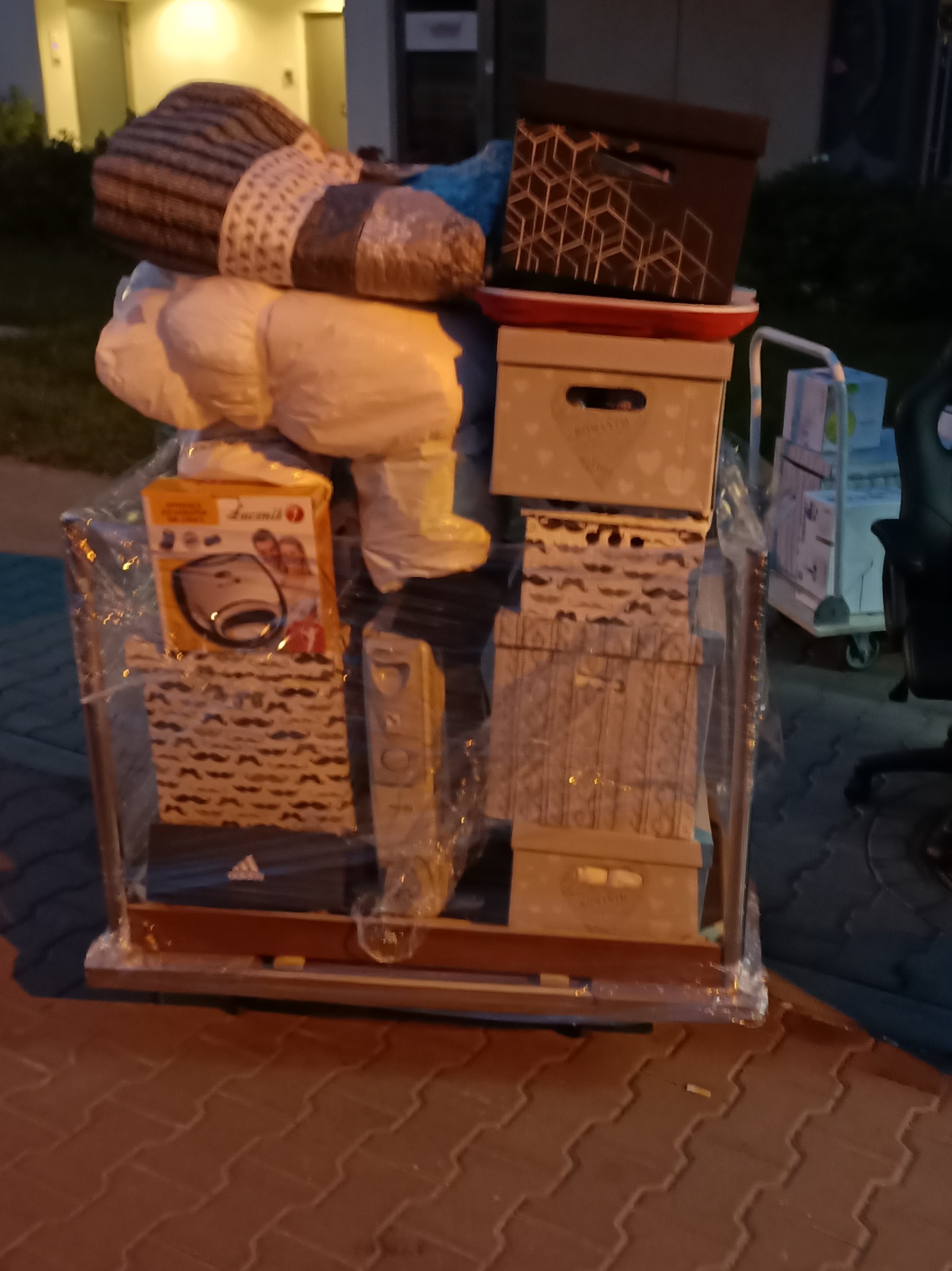 Wózek transportowy załadowany kartonami, workami i sprzętem AGD, zabezpieczony folią stretch, na chodniku z kostki brukowej.