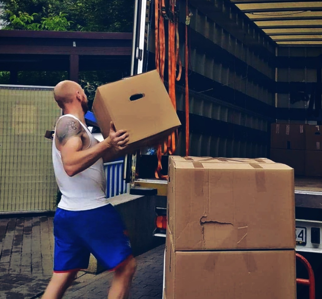 Mężczyzna z tatuażem na ramieniu w białym podkoszulku i niebieskich spodenkach przenosi kartonowe pudło do wnętrza ciężarówki, widoczne pasy transportowe i inne kartony.