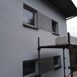 Kompleksowe remonty i elewacje - Fragment elewacji domu z oknami, częściowo otynkowany fragment ściany z widoczną fakturą, rusztowanie z bloczkiem styropianu na górze, pochmurne niebo.