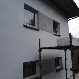 Fragment elewacji domu z oknami, częściowo otynkowany fragment ściany z widoczną fakturą, rusztowanie z bloczkiem styropianu na górze, pochmurne niebo.