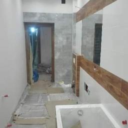 Kompleksowe remonty i elewacje - Wnętrze łazienki w trakcie remontu, zabezpieczona podłoga kartonem, częściowo wykończone ściany z białymi i drewnopodobnymi płytkami, widoczna wanna i przygotowane miejsce na lustro.