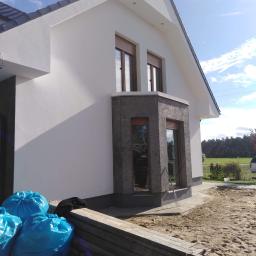 Kompleksowe remonty i elewacje - Nowoczesny dom jednorodzinny z białą elewacją i ciemnoszarą wstawką okienną, widoczne worki z odpadami budowlanymi i drewniane stemple szalunkowe na placu budowy.