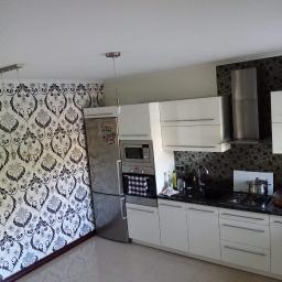 Kompleksowe remonty i elewacje - Jasna kuchnia z białymi szafkami, czarnym blatem i wzorzystą tapetą na ścianach, widok na zabudowę kuchenną i podłogę z płytek.