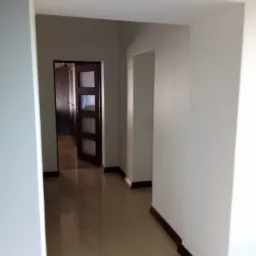 Korytarz z jasnymi, świeżo pomalowanymi ścianami, brązowymi listwami przypodłogowymi i błyszczącą, beżową podłogą. W głębi widoczne drzwi z szybami i inne pomieszczenia.