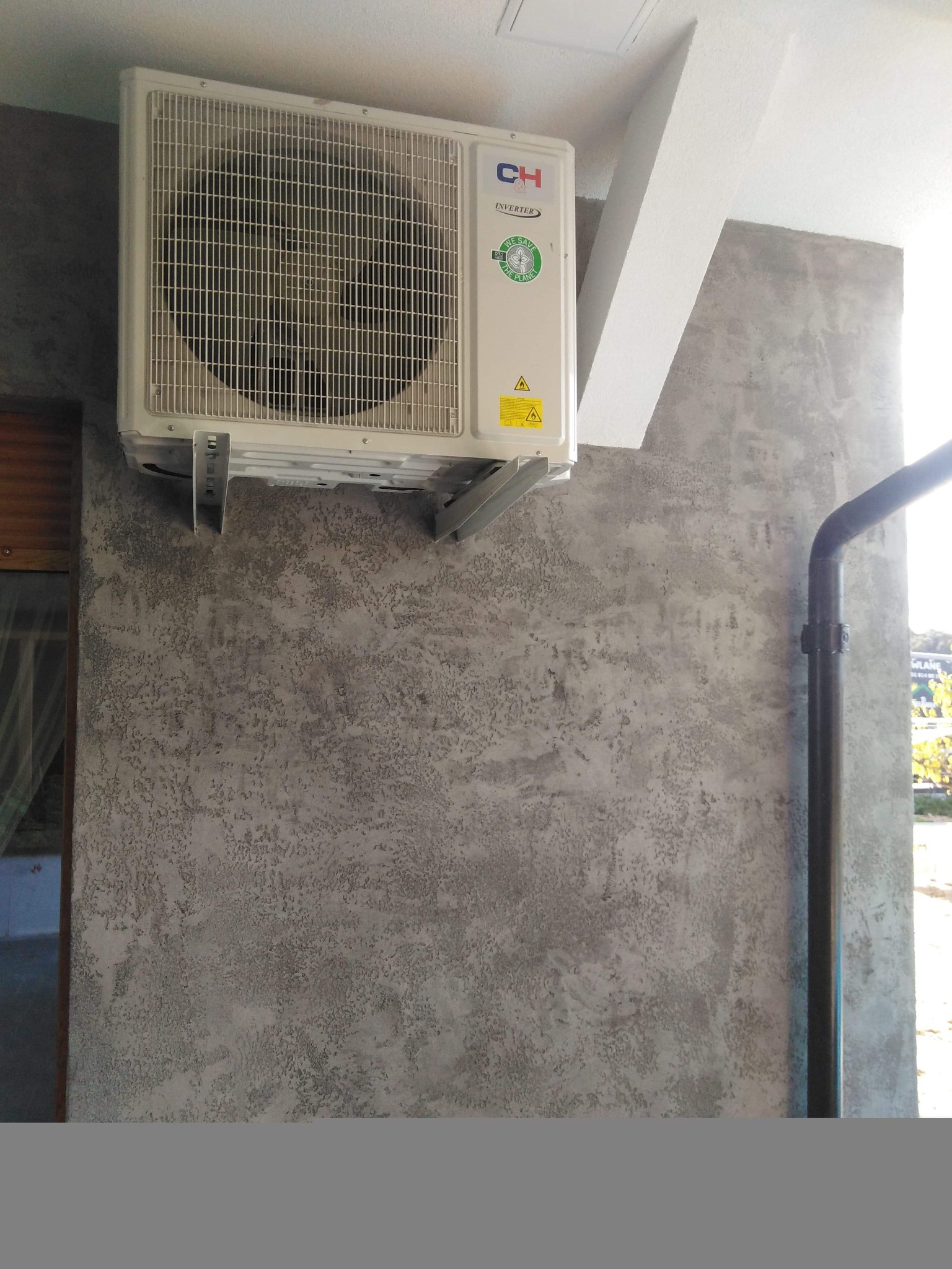 Zamontowany klimatyzator marki CH Inverter na szarej, teksturowanej ścianie zewnętrznej budynku z widoczną rurą spustową i fragmentem okna.