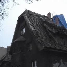 Stary, ciemny budynek z uszkodzonym dachem pokrytym dachówką, zabezpieczonym niebieską plandeką i żółtym zsypem na gruz.