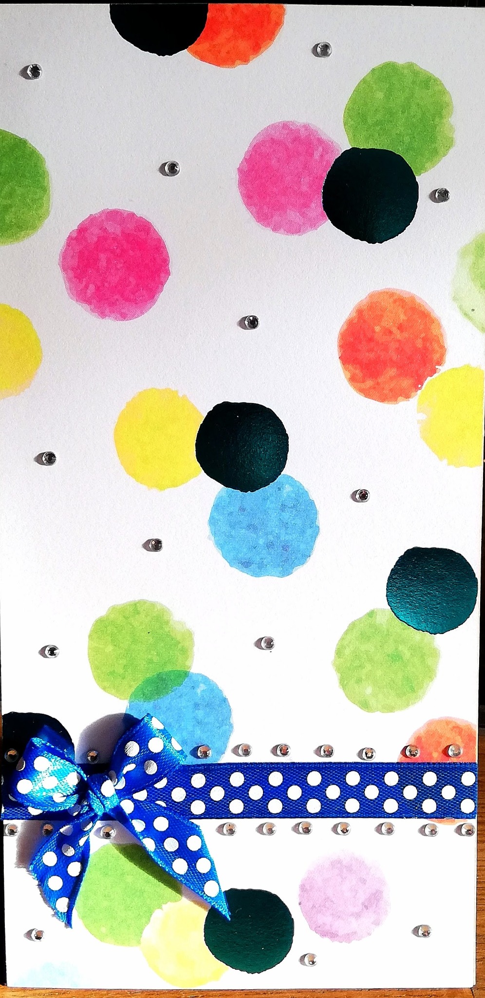Ręcznie robiona kartka imieninowa w stylu scrapbookingu, z akwarelowymi kropkami w kolorach tęczy, ozdobiona niebieską wstążką w białe groszki i srebrnymi dżetami.