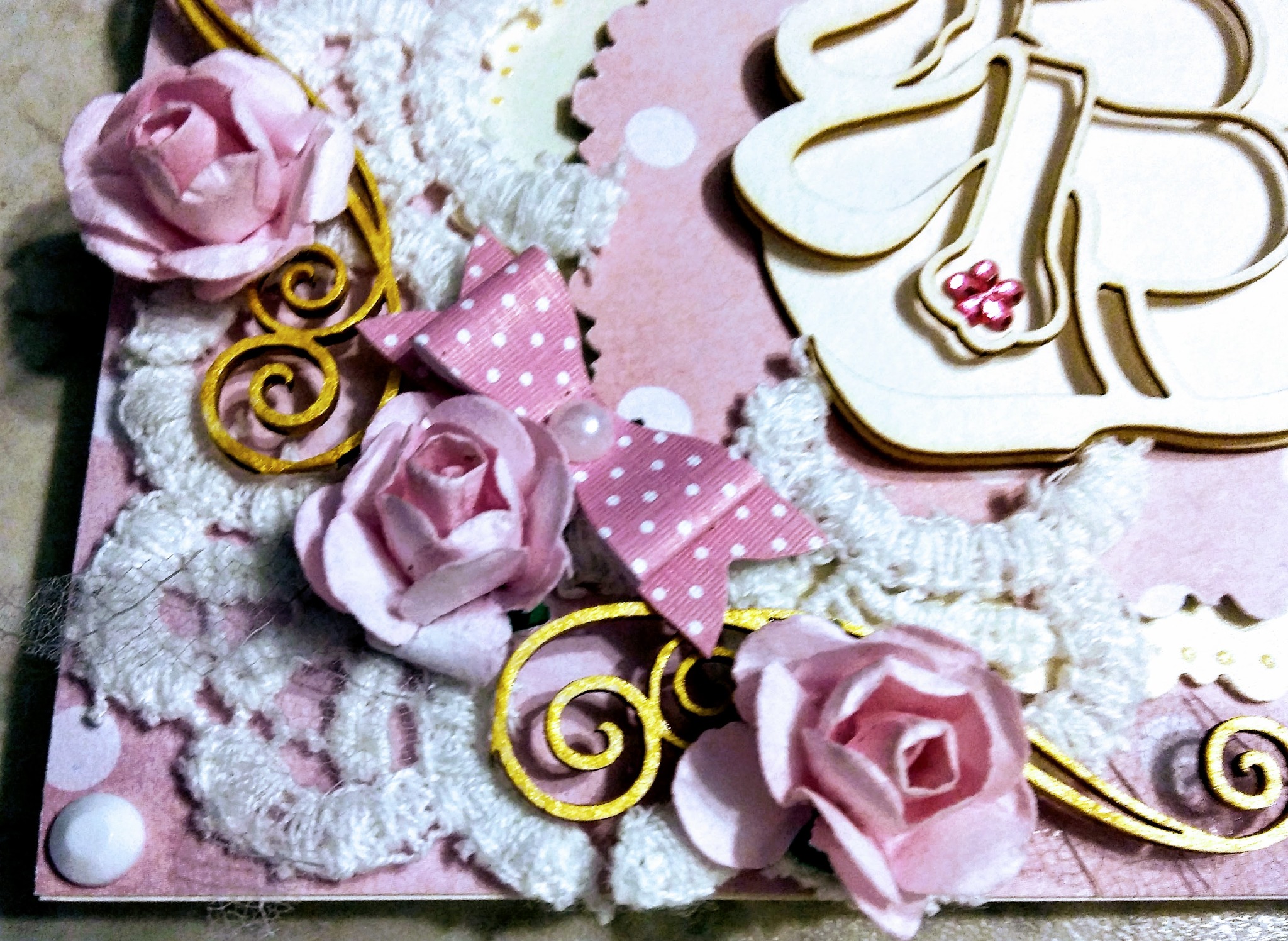 Ręcznie robiona kartka z okazji urodzin w stylu scrapbookingu, zdobiona różowymi papierowymi różami, koronką, kokardką w białe kropki i złotymi ornamentami na różowym tle.