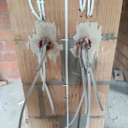 Fragment ściany z cegły dziurawki z zamontowanymi okrągłymi puszkami instalacyjnymi i wyprowadzonymi przewodami elektrycznymi, na których widoczne są odręczne napisy.