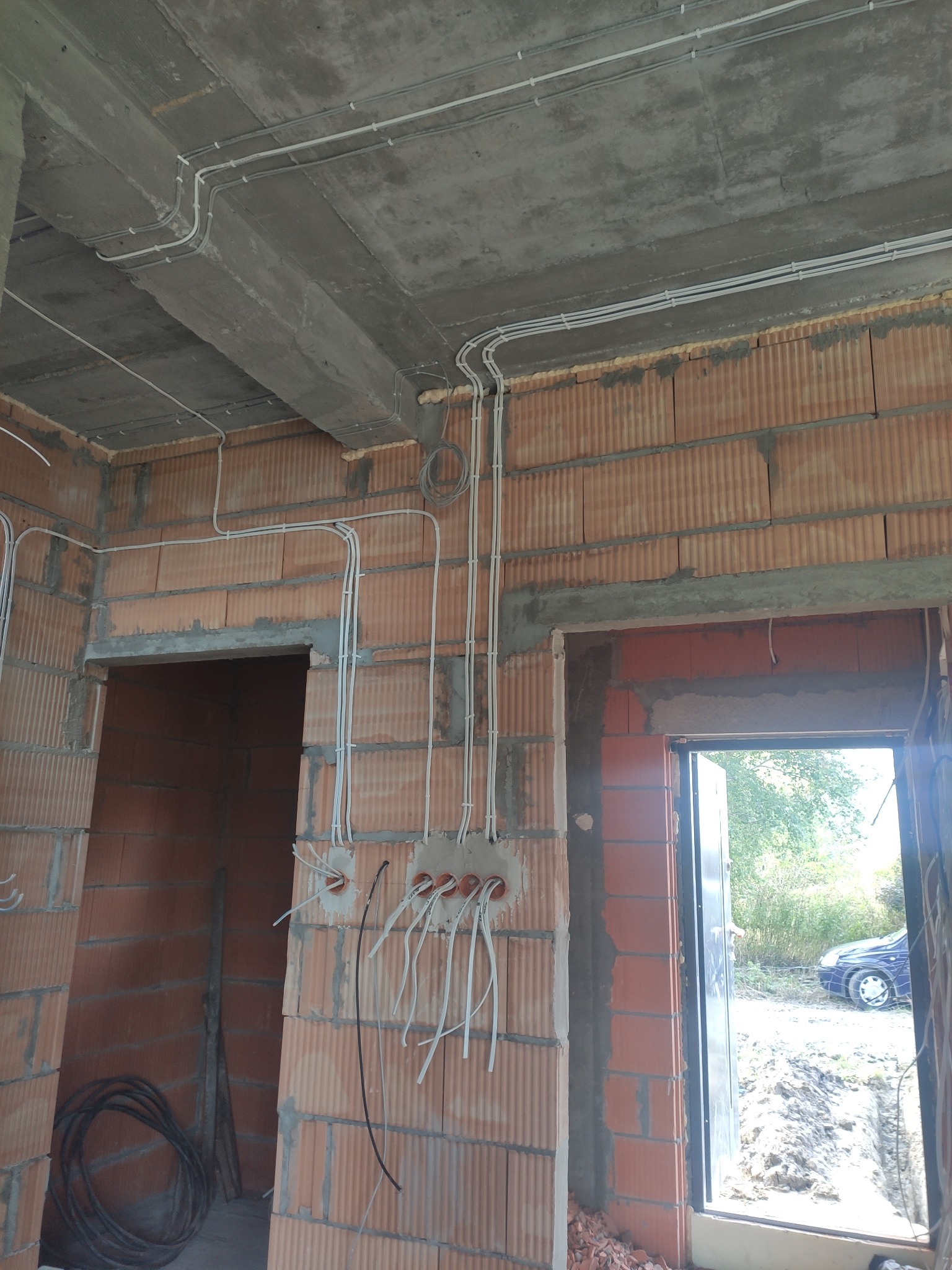 Wewnętrzne rozprowadzenie instalacji elektrycznej w surowym budynku z cegły, widoczne kable prowadzone po suficie i ścianach, puszki instalacyjne z wystającymi przewodami.