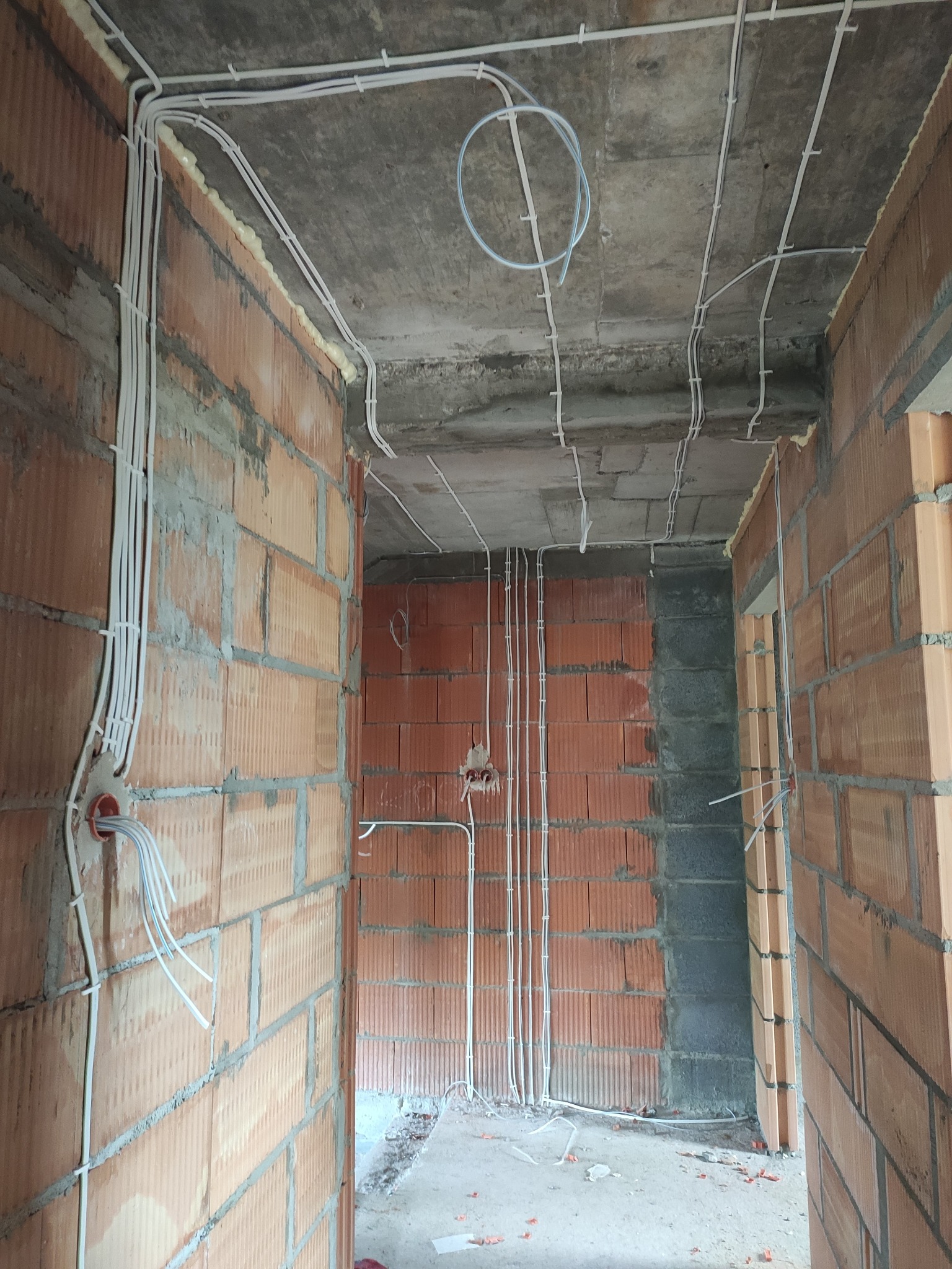 Wnętrze w trakcie budowy z widocznymi przewodami elektrycznymi poprowadzonymi po ścianach z cegły i betonowym suficie. Kable przymocowane do sufitu za pomocą uchwytów, a pianka montażowa uszczelnia...