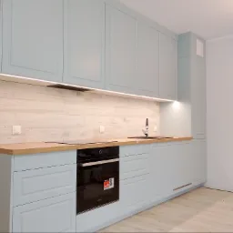 Jasna, błękitna zabudowa kuchenna z drewnianym blatem i płytkami imitującymi drewno na ścianie. Nowoczesny design z oświetleniem LED podszafkowym i czarnym zlewem.