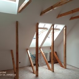 Zakład usługowy Piotr Pionk - Poddasze w trakcie adaptacji: drewniane belki wzmacniające konstrukcję dachu, surowe ściany przygotowane do ocieplenia, widoczne okna dachowe i fragment grzejnika.