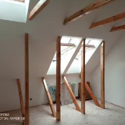 Poddasze w trakcie adaptacji: drewniane belki wzmacniające konstrukcję dachu, surowe ściany przygotowane do ocieplenia, widoczne okna dachowe i fragment grzejnika.