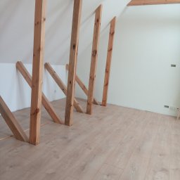 Zakład usługowy Piotr Pionk - Poddasze po montażu drewnianej konstrukcji nośnej, widoczne ukośne słupy i fragmenty belek stropowych, świeżo pomalowane ściany na biało, podłoga wyłożona panelami w odcieniach beżu.