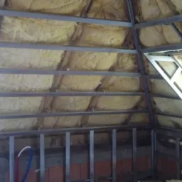 Konstrukcja metalowa pod płyty gipsowo-kartonowe na poddaszu z oknem dachowym, ocieplona wełną mineralną, widoczne fragmenty instalacji wodnej i muru z cegły.