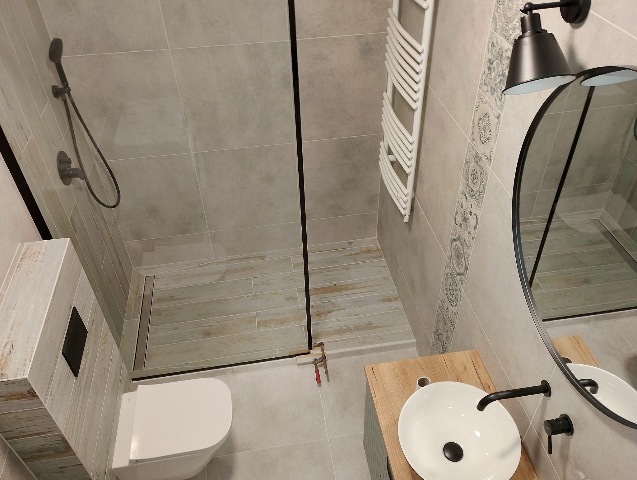 Nowoczesna łazienka z kabiną typu walk-in, drewnopodobnymi płytkami na podłodze prysznica i okrągłym lustrem z czarnymi akcentami. Widok z góry.