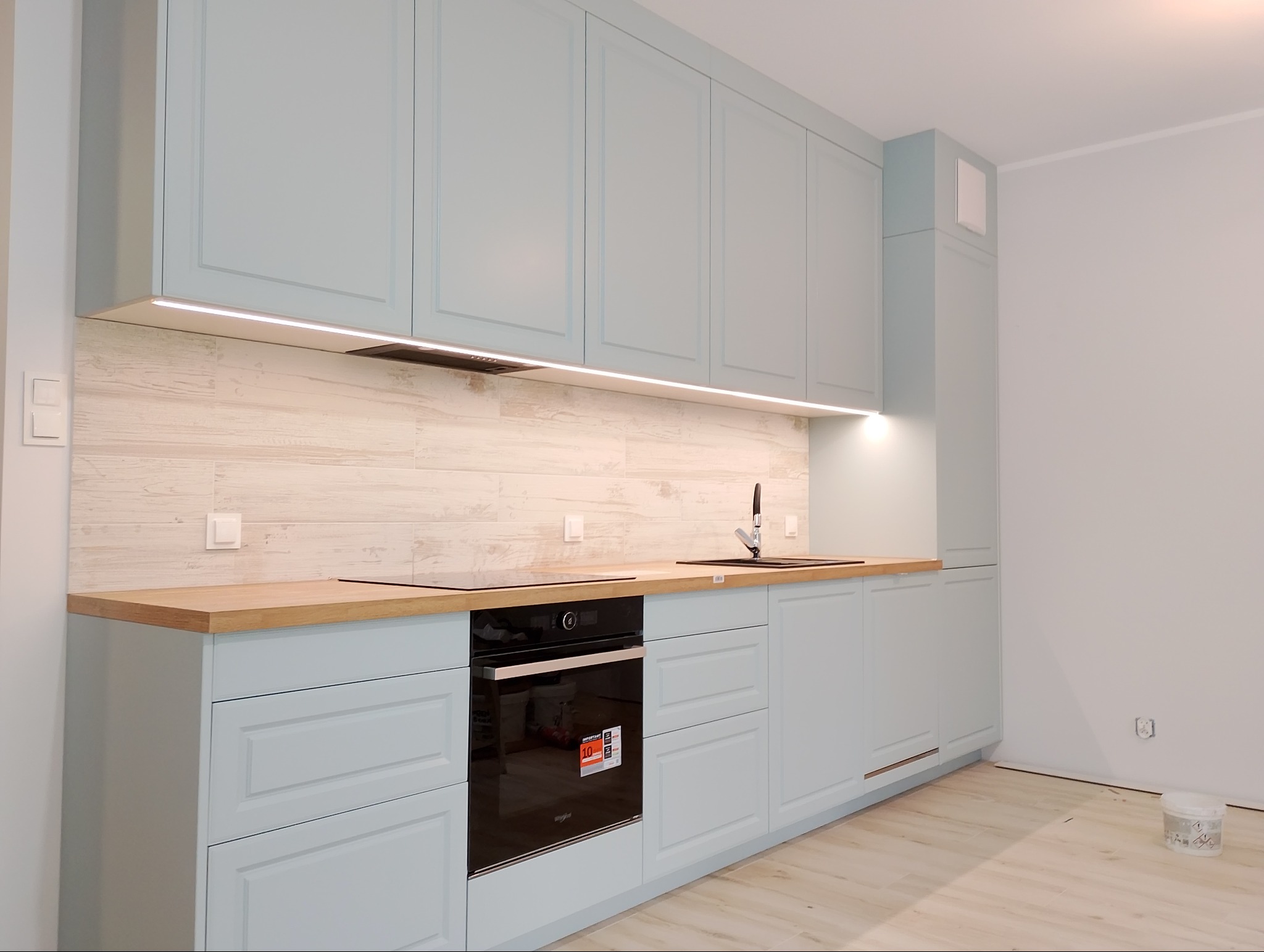 Jasna, błękitna zabudowa kuchenna z drewnianym blatem i płytkami imitującymi drewno na ścianie. Nowoczesny design z oświetleniem LED podszafkowym i czarnym zlewem.