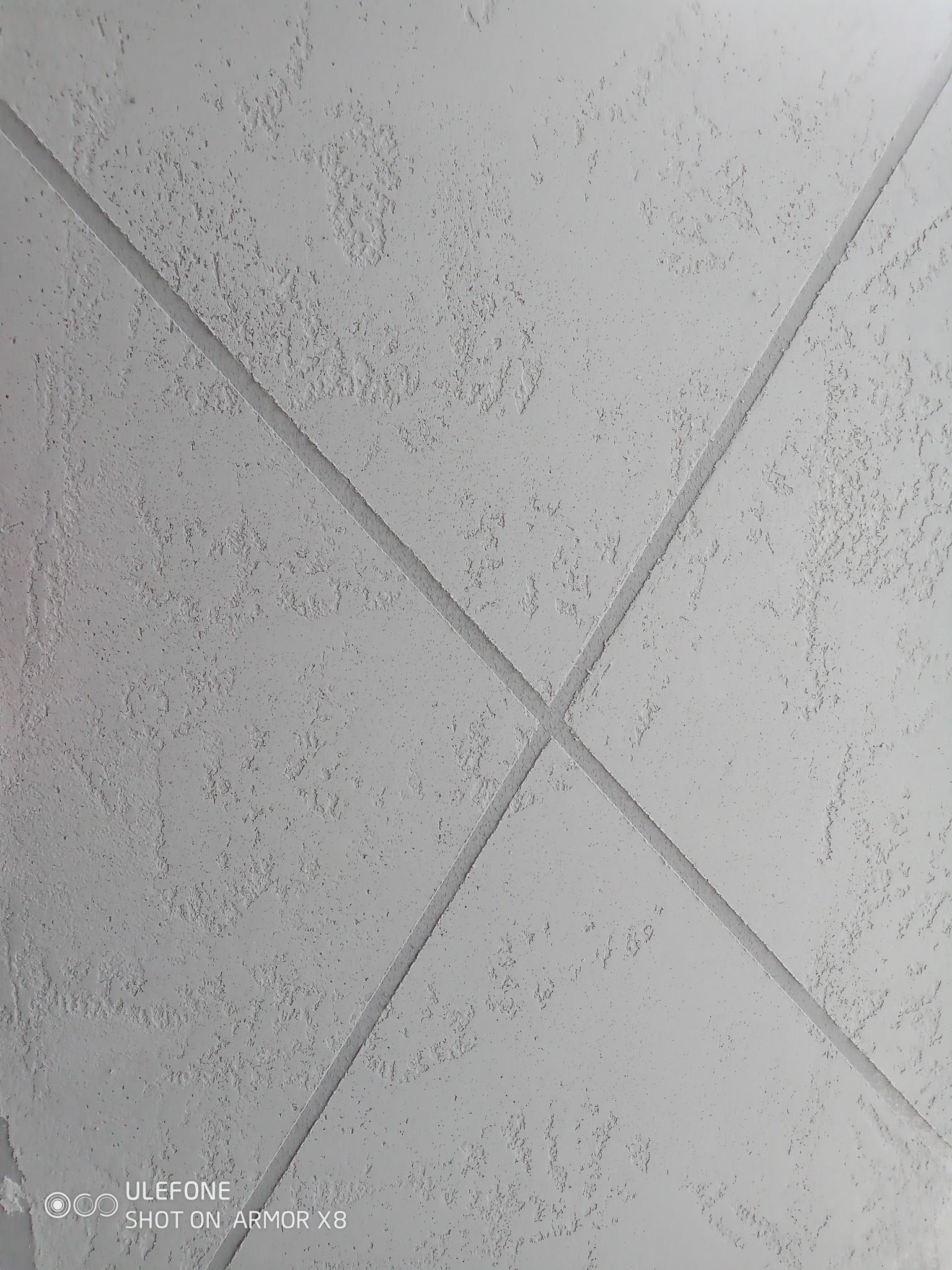 Szara ściana z fakturą i wyżłobionymi liniami tworzącymi wzór rombów, wykonana z bliska, z widoczną teksturą tynku.