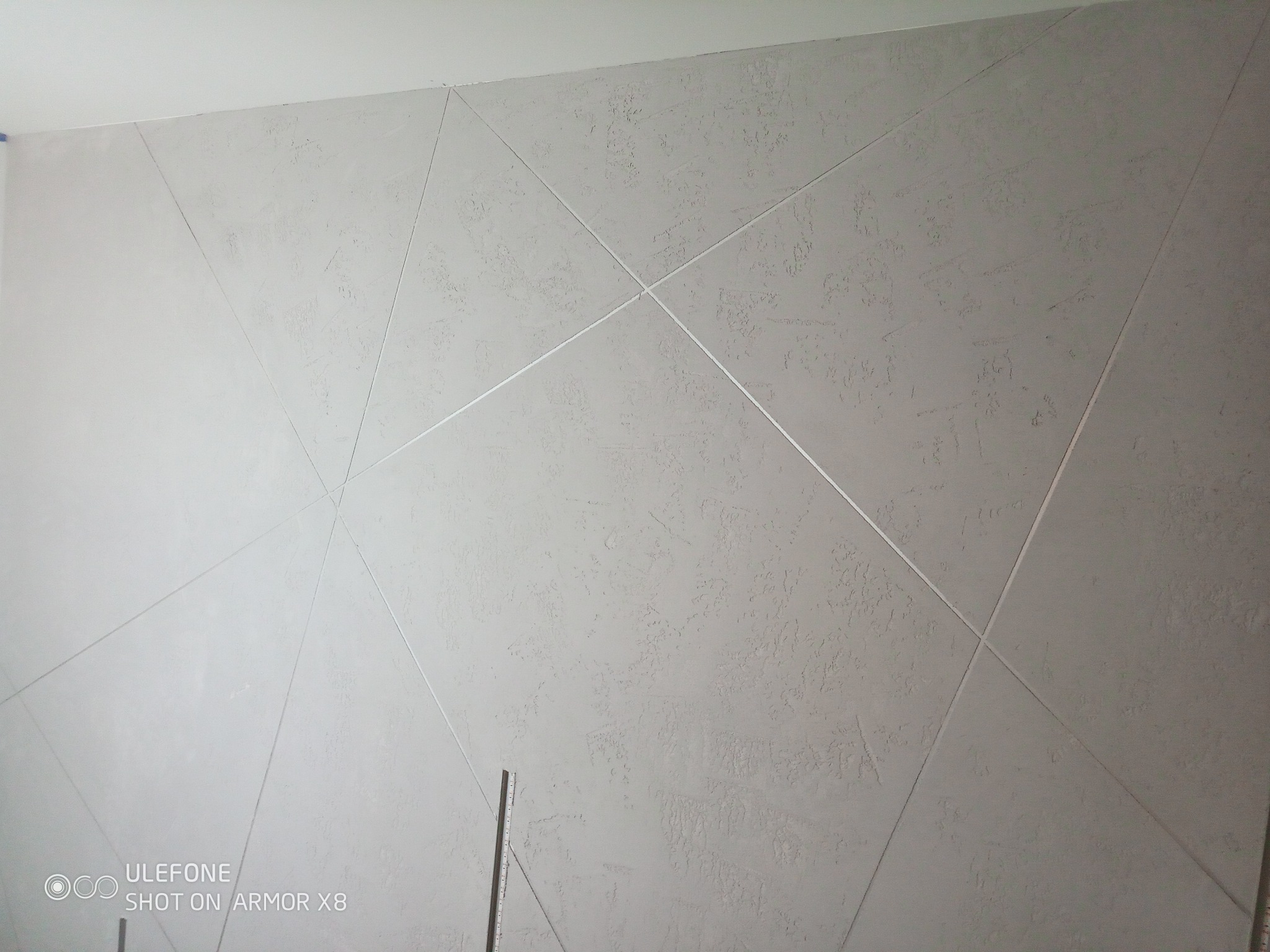 Teksturowana ściana w odcieniach szarości z geometrycznym wzorem z delikatnych linii, widoczny fragment listwy przypodłogowej w dolnej części kadru.