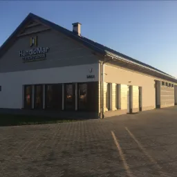 Budynek firmy HandloMar Centrum z napisem na elewacji i stosem palet w tle, oświetlony popołudniowym słońcem