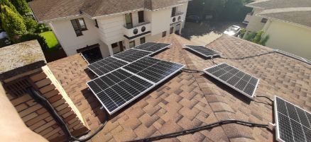 Panele słoneczne zamontowane na dachu domu z brązową dachówką bitumiczną, widoczny komin z cegły i fragment elewacji budynku.