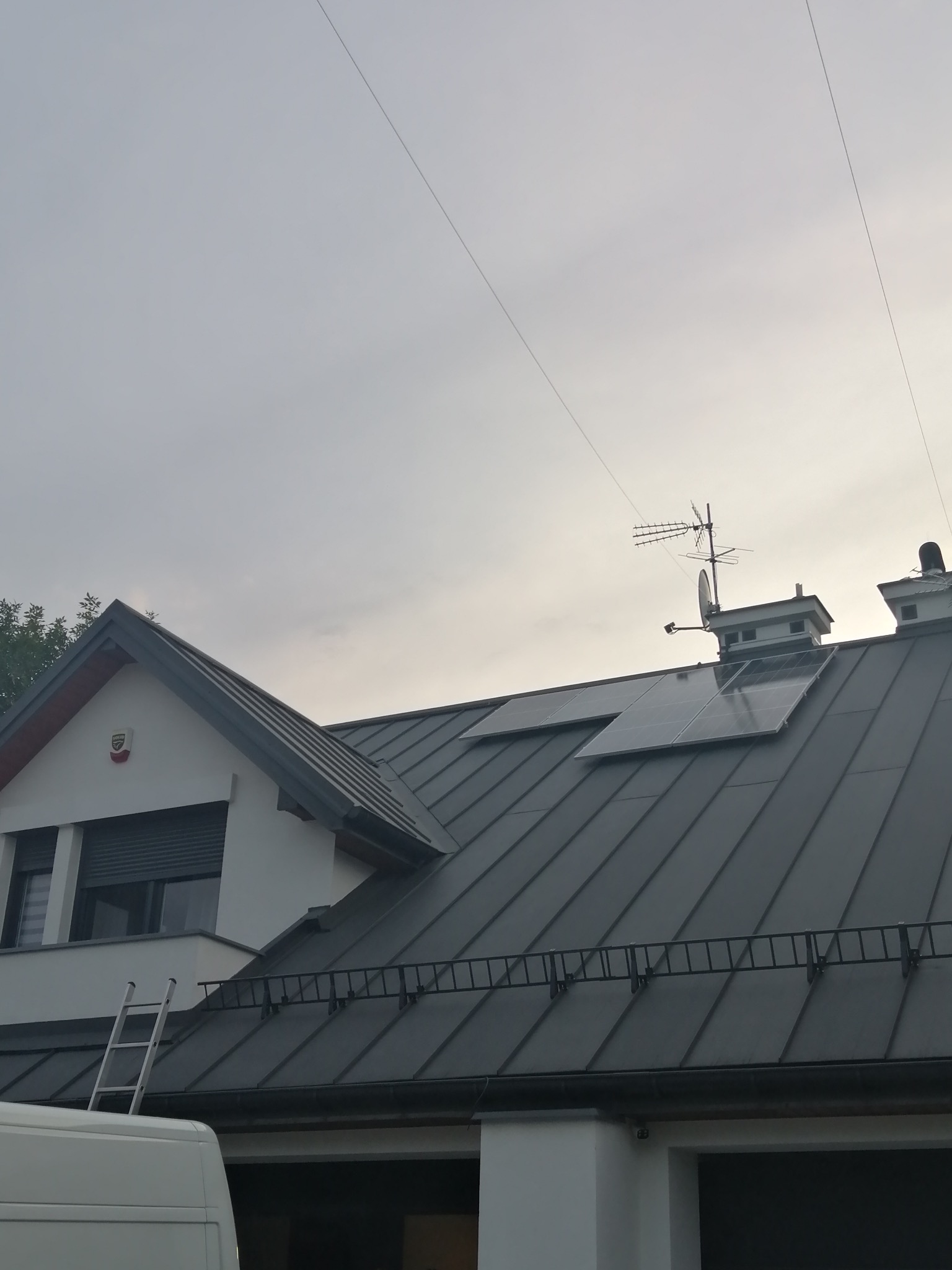 Panele słoneczne na metalowym dachu domu jednorodzinnego z anteną telewizyjną, drabina oparta o budynek, widoczny fragment białego samochodu dostawczego.