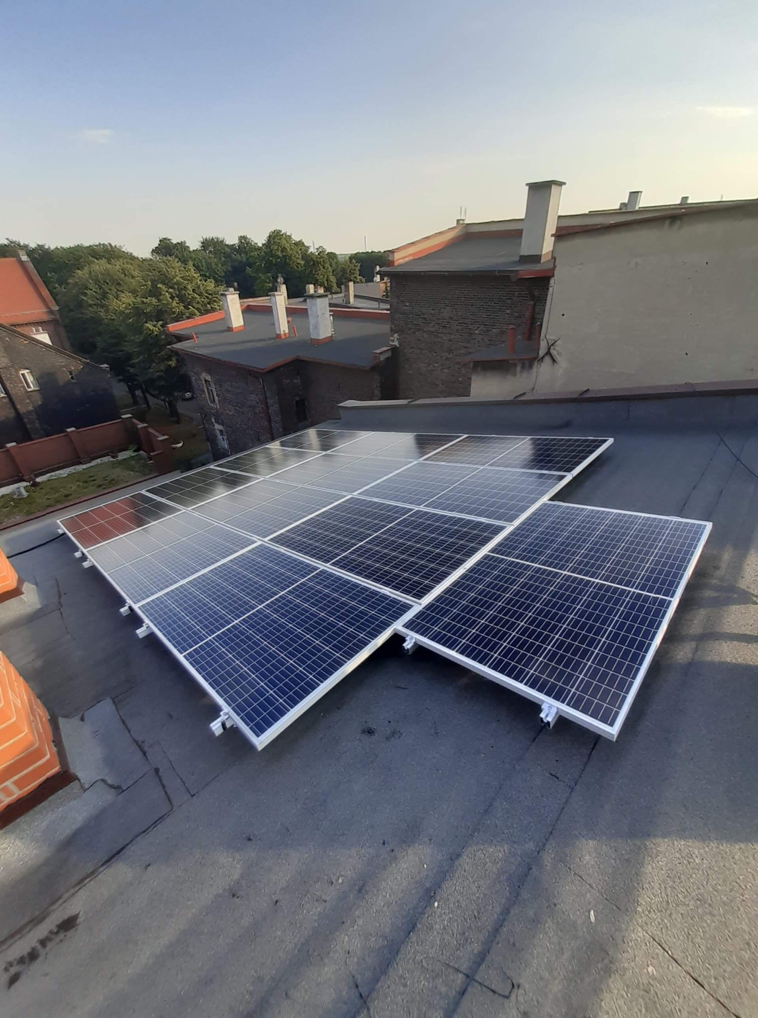 Panele słoneczne zamontowane na płaskim dachu budynku mieszkalnego, widok z góry, w tle budynki z cegły i drzewa.