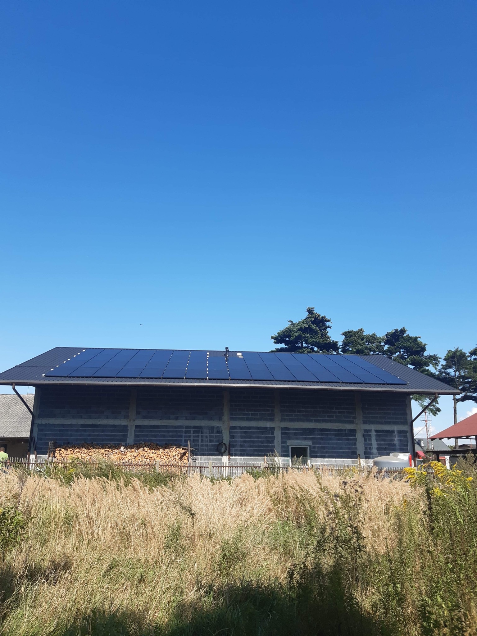 Niedokończony budynek gospodarczy z panelami słonecznymi na dachu, stos drewna opałowego, wysoka trawa na pierwszym planie, błękitne niebo.